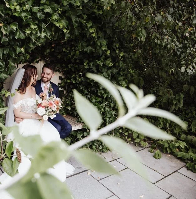 Bride and groom sitting on a bench surrounded by greenery, bride holding a bouquet.