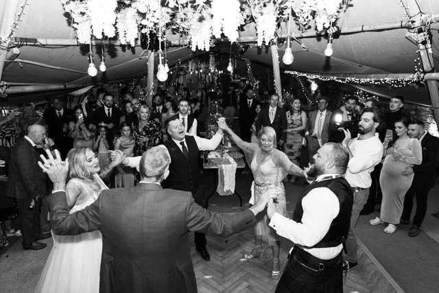 A black and white wedding reception scene featuring a group of people dancing in a circle. The event is held under a decorated tent, with floral garlands and string lights hanging overhead. Guests are standing around, watching and enjoying the festivities. The atmosphere is lively and celebratory.