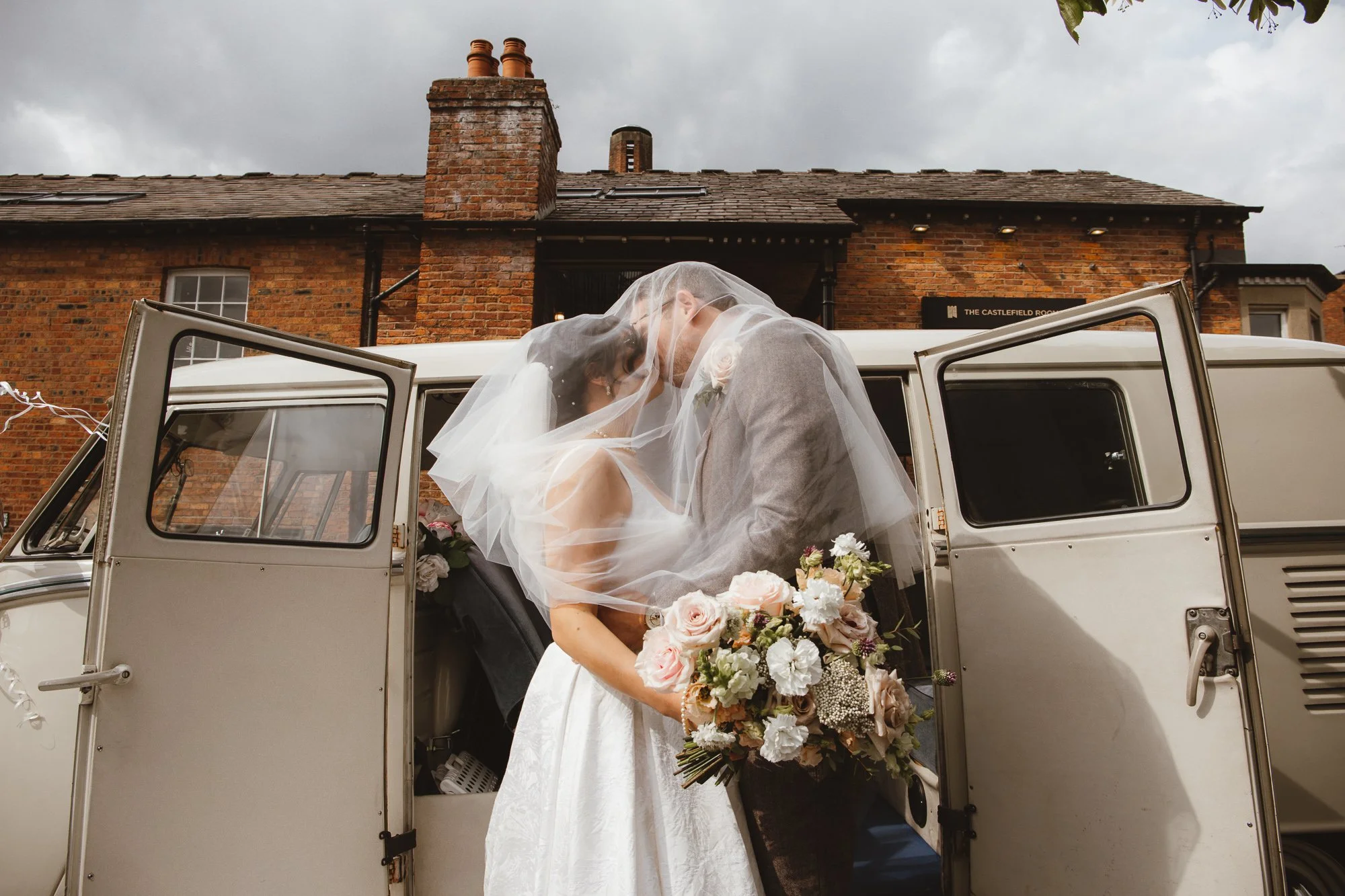 A bride and groom share a kiss inside a vintage car, with the bride holding a bouquet, in front of a brick building on a cloudy day.