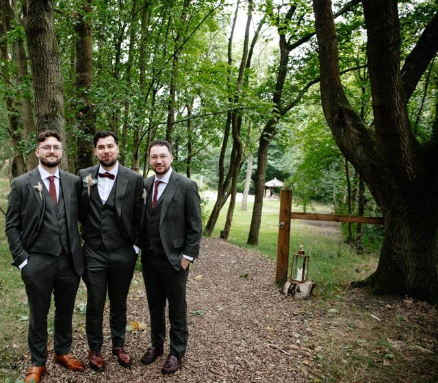 Three men in suits and ties posing in a wooded outdoor setting.
