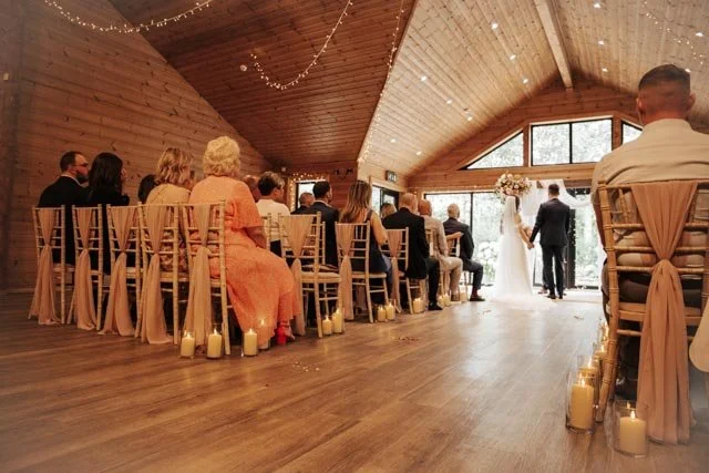 A rustic indoor wedding ceremony in a wooden hall, with guests seated on decorated chairs and the bride and groom standing at the end of the aisle. Candles line the aisle, and string lights adorn the ceiling.
