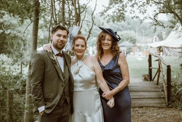 Three people pose outdoors on a path in formal attire; one man in a suit and two women in elegant dresses, standing near a small wooden bridge surrounded by trees.
