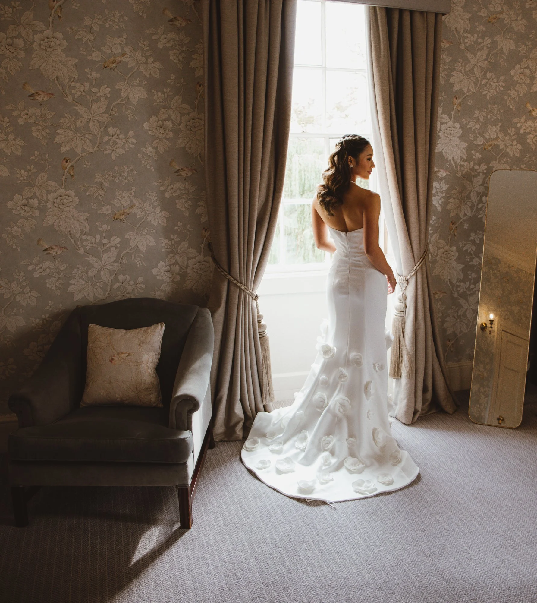 A Bride in a white wedding dress standing by a window, looking outside in a room with floral wallpaper, beige curtains, a gray armchair with a patterned pillow, and a full-length mirror.