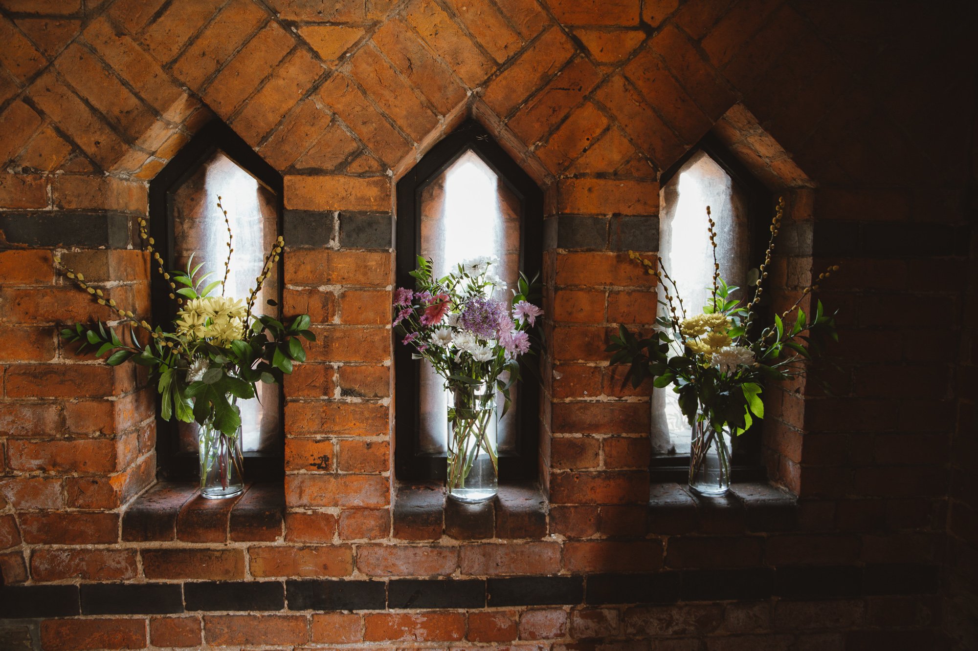 Three glass vases with flower arrangements on a brick ledge below three small arched stained glass windows in a brick wall.