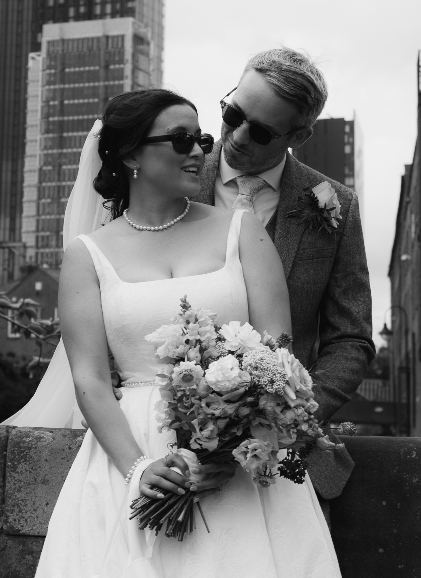 Black and white photo of a bride and groom looking at each other, with the bride holding a large bouquet of flowers. They are standing outdoors with city buildings in the background, both wearing sunglasses.