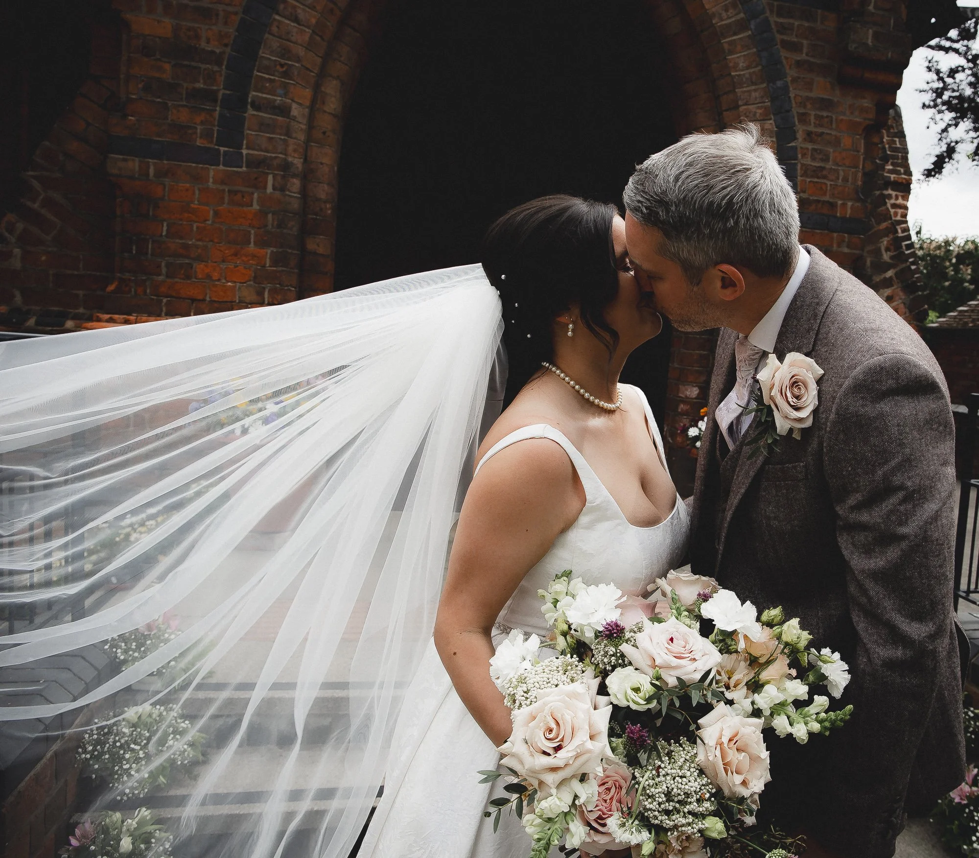 A bride and groom kissing outdoors, with the bride wearing a white dress, pearl necklace, and veil, holding a bouquet of pink and white flowers, and the groom in a gray suit with a boutonniere, standing in front of a brick structure.