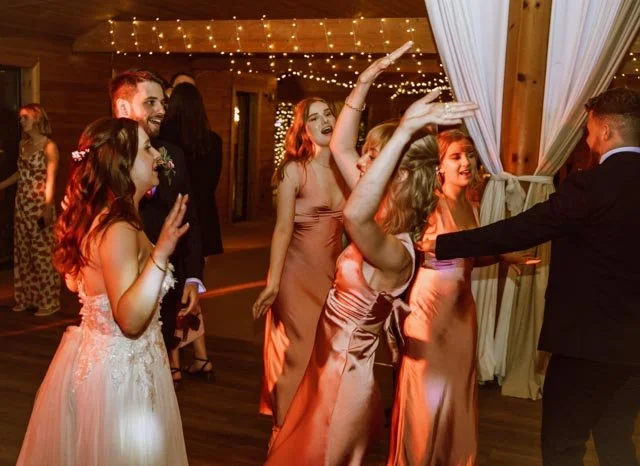 Group of people dancing at a wedding reception, women in formal dresses, and fairy lights in the background.