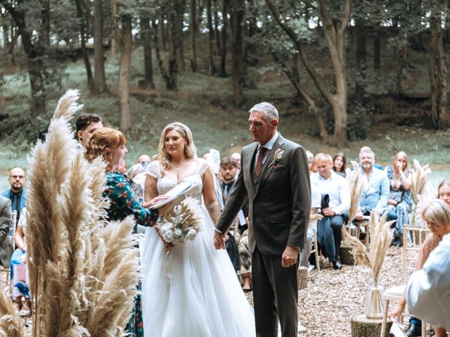 Outdoor wedding ceremony in a forest setting with a bride in a white gown and a groom in a suit holding hands, officiated by a woman. Guests are seated, surrounded by decorative pampas grass.