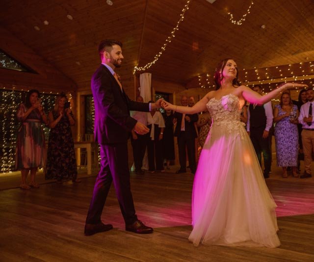 Bride and groom dancing at a wedding reception with guests watching, set in a warmly lit venue with string lights.