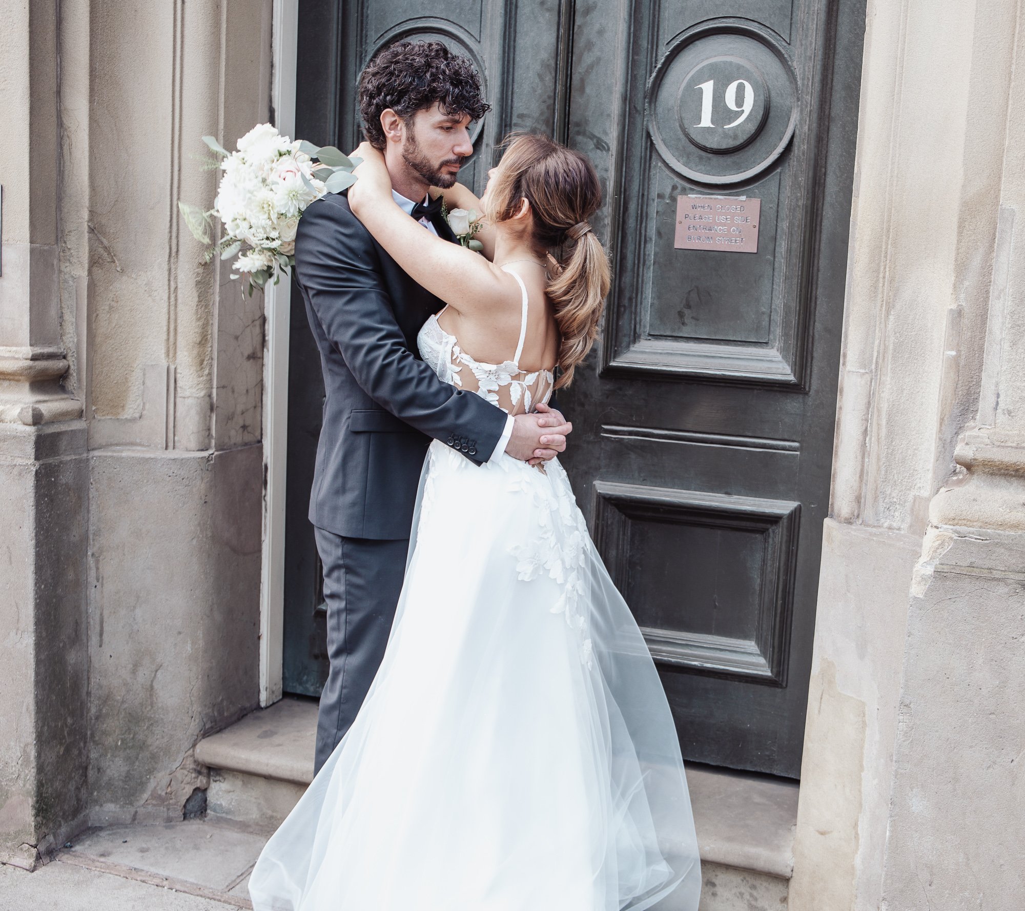 Bride and groom embracing in front of a dark door with number 19; bride in white gown, groom in dark suit, holding a bouquet of white flowers.
