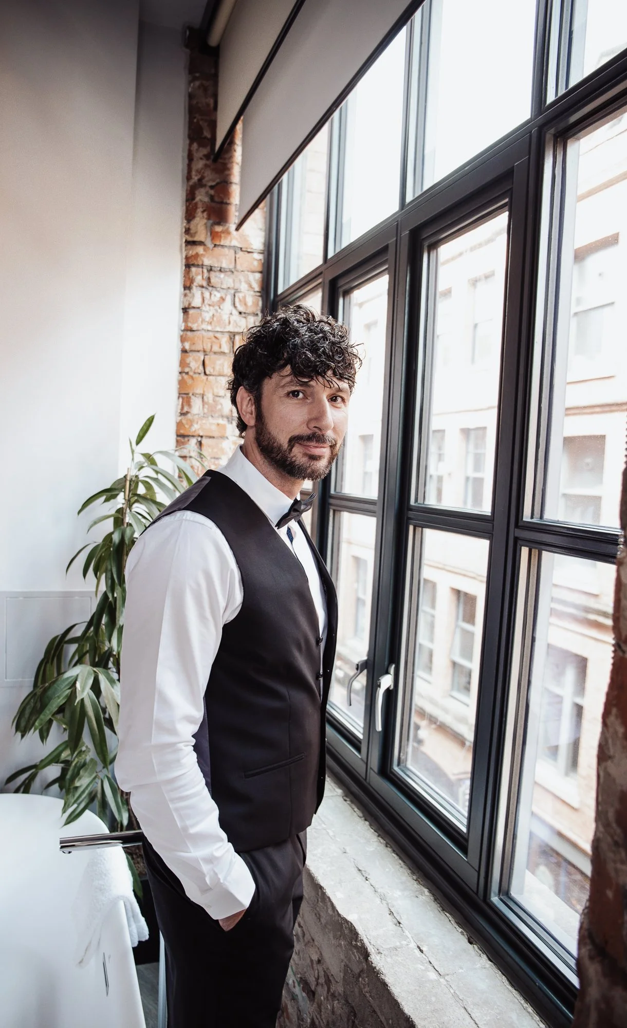 A man with curly hair and a beard wearing a black vest, white shirt, and bow tie standing by a window in a room with exposed brick walls and a potted plant.