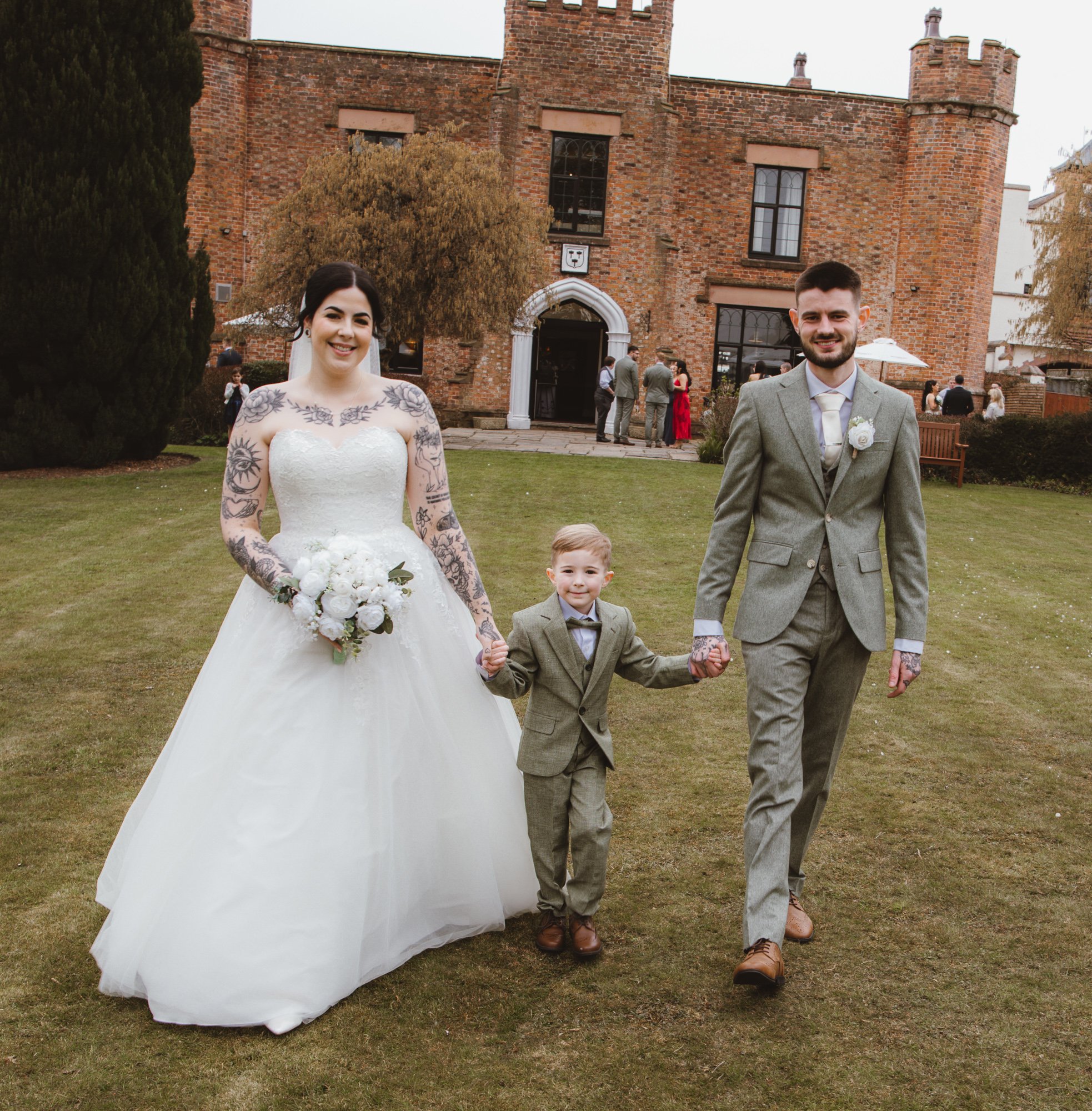 A wedding couple and a young boy walking on grass in front of a brick building. The bride is wearing a white gown and holding a bouquet, and the groom and boy are in matching gray suits.