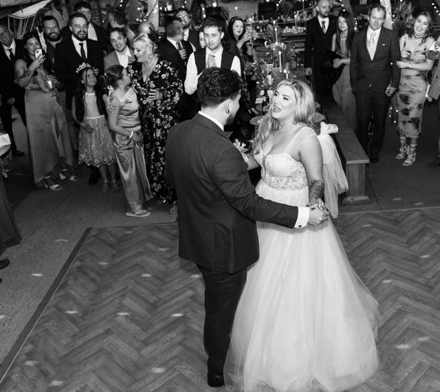 A bride and groom dancing at their wedding reception, surrounded by guests watching and smiling. The scene is festive, with people wearing formal attire.