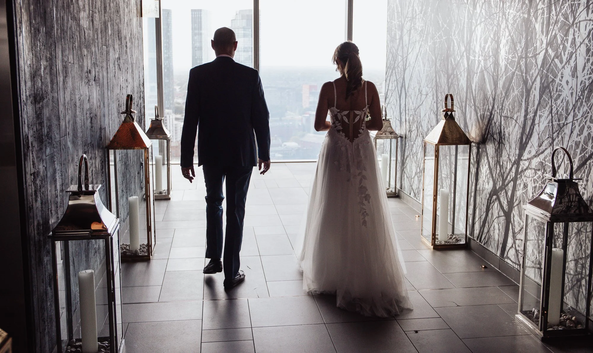 Silhouette of a woman in a wedding dress and a man walking down a corridor with large windows and decorative lanterns.