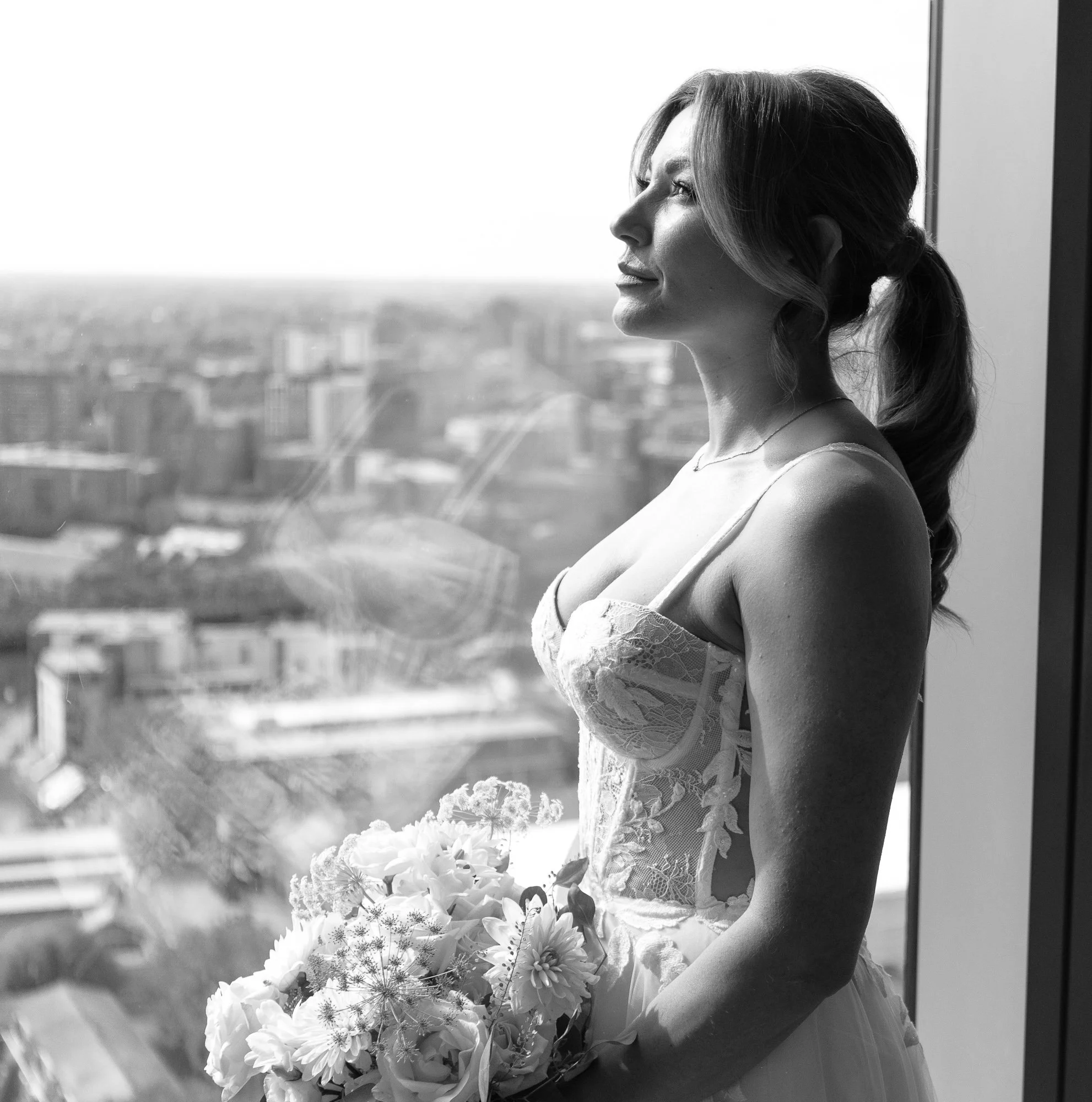 Bride holding a bouquet of flowers, standing by a window overlooking a cityscape, wearing a lace wedding dress; black and white photograph.