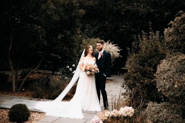 Bride and groom in a garden setting, smiling and holding a bouquet, with greenery and flowers around them.