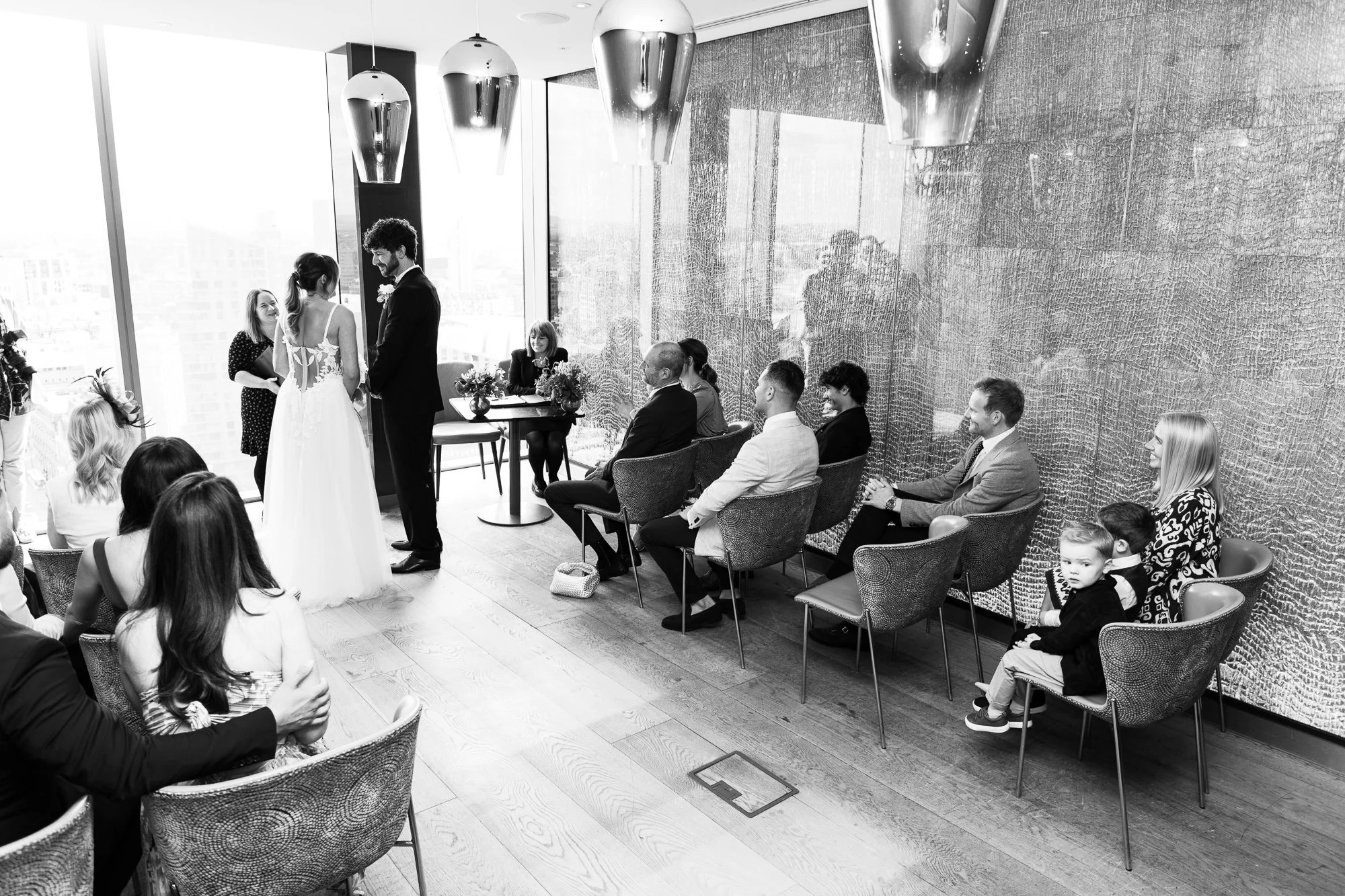 Black and white photo of an indoor wedding ceremony with a couple standing and an officiant in front of seated guests in a modern setting.