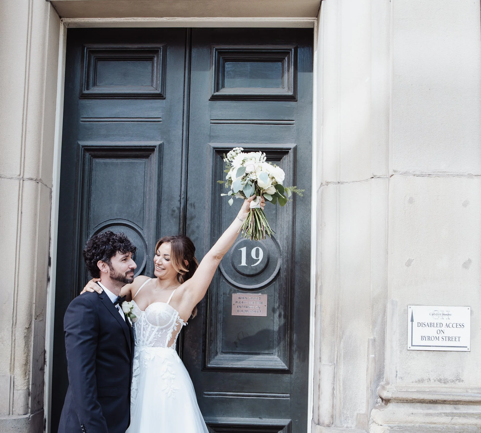 Bride and groom posing in front of large door with number 19, bride holding bouquet.