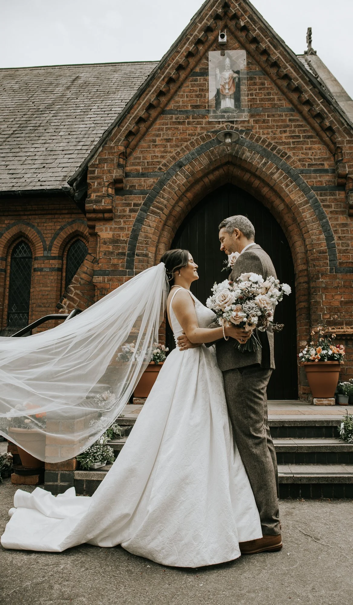 A bride and groom standing close to each other outside a brick church, holding a bouquet of flowers, smiling, and looking into each other's eyes on their wedding day.