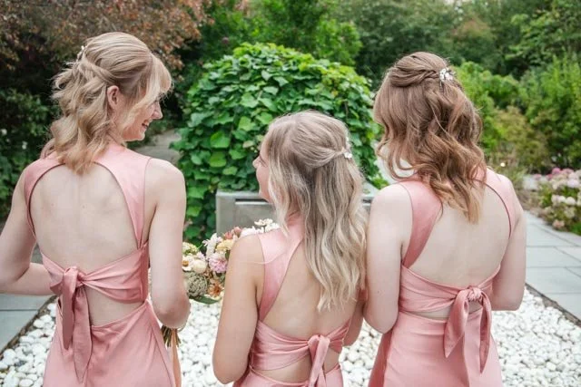 Three women in matching pink dresses standing outdoors, with their backs facing the camera. They are in a garden setting with greenery and stones.
