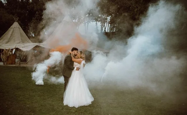 Bride and groom kissing outdoors in front of lit tents with smoke surrounding them.
