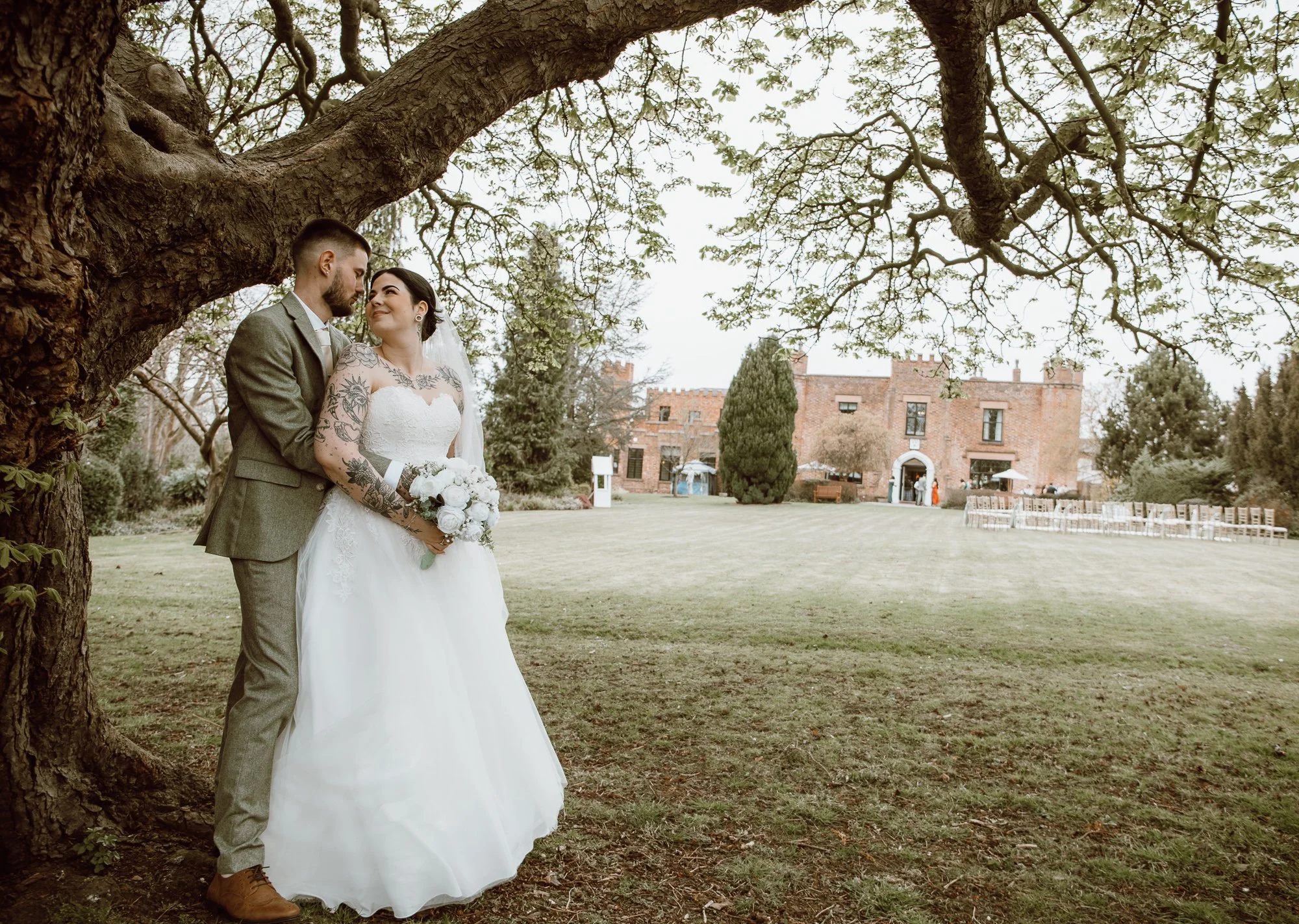 Bride and groom standing under a tree in a garden with a historic building in the background.