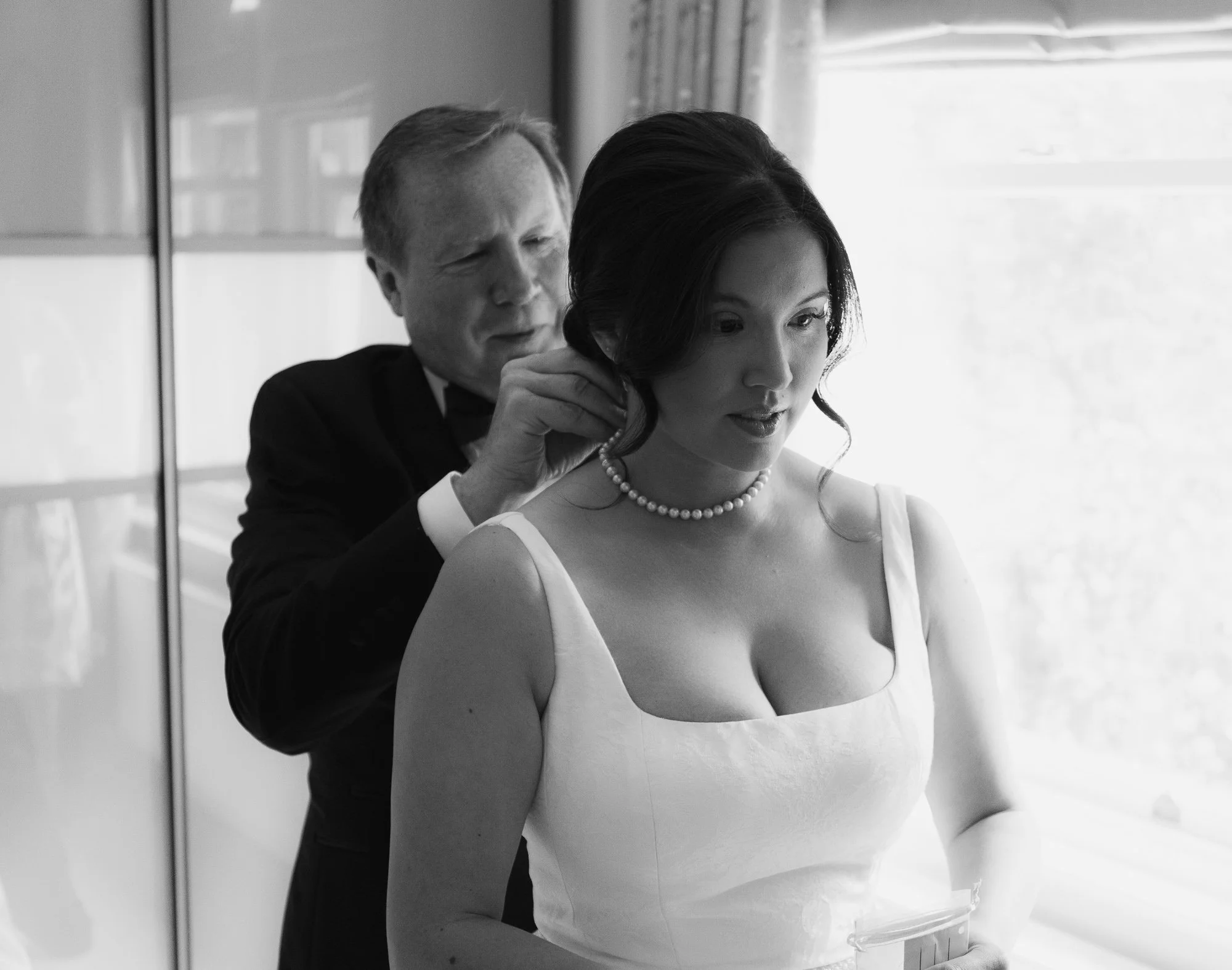 A man helping a woman get ready, dressed in a white gown with a pearl necklace, in a well-lit room.