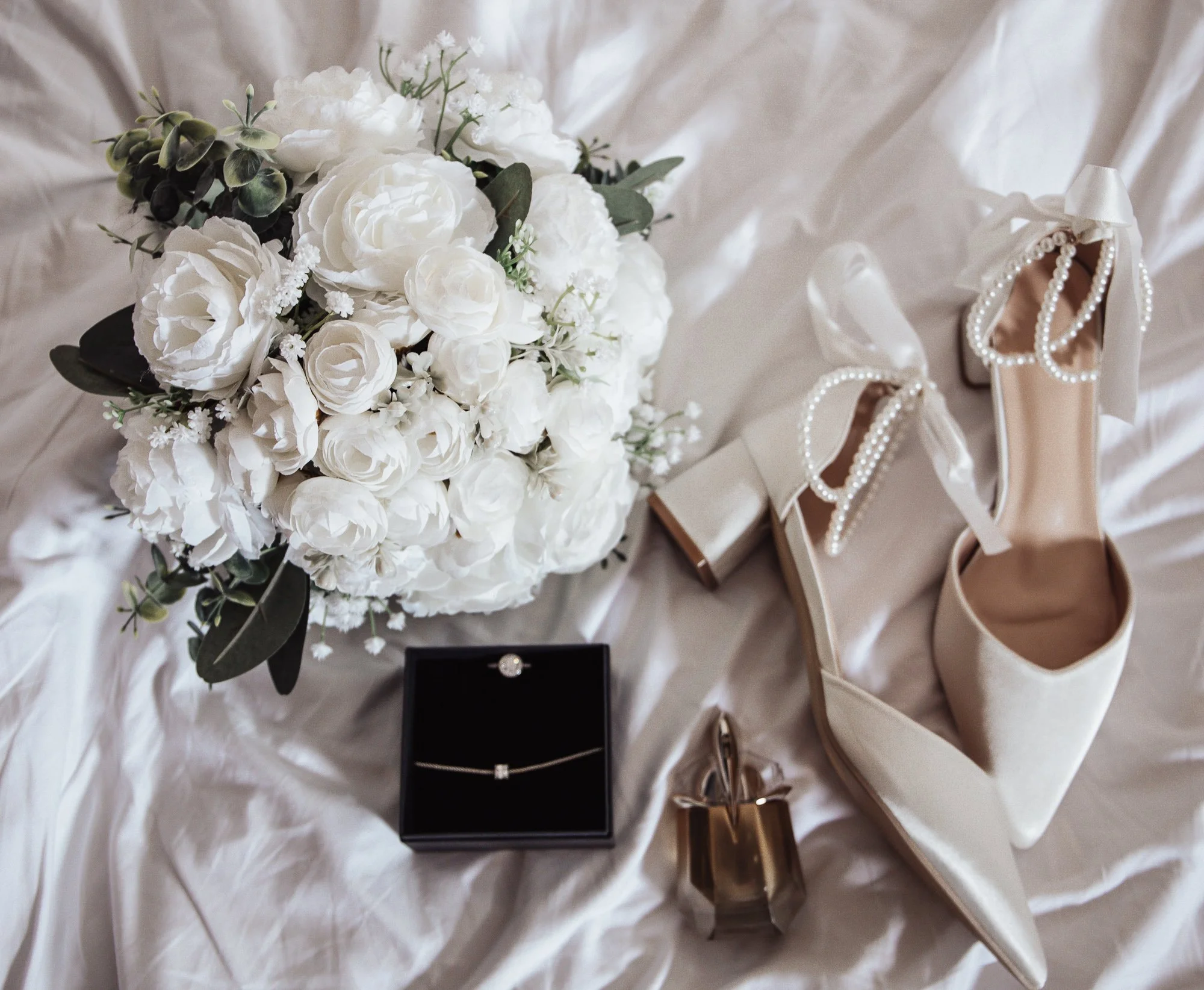 Bridal flat lay with white bouquet, pearl-embellished shoes, jewelry box, and perfume bottle on white fabric.