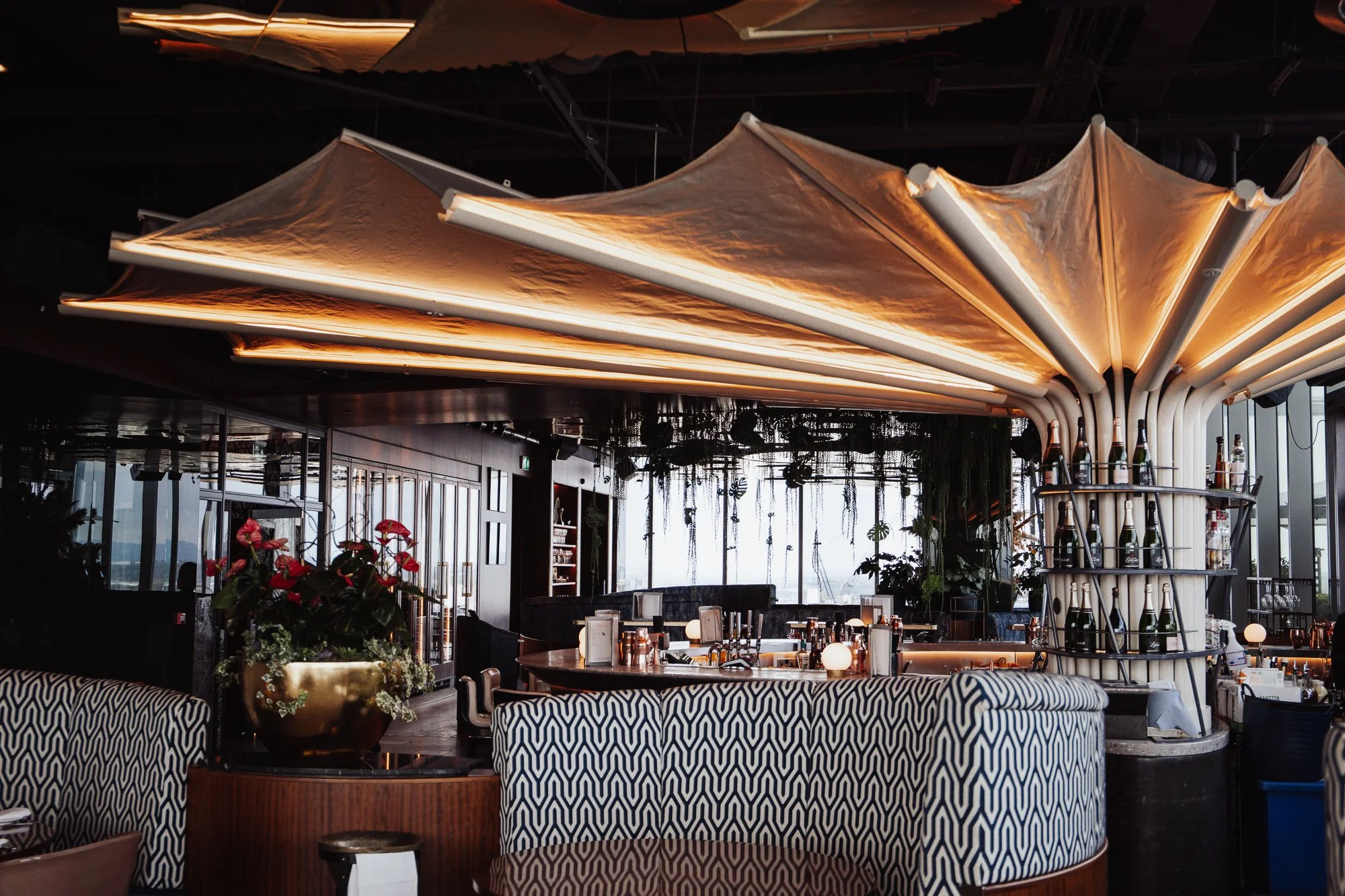 Interior of a modern bar with patterned seating, decorative lighting, plants, and shelves filled with bottles, creating an elegant ambiance.