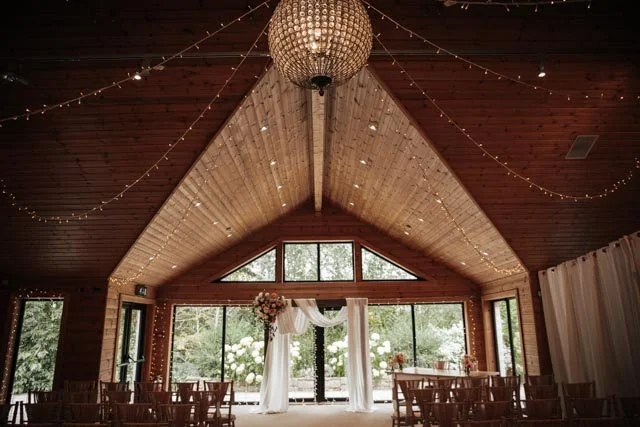 Interior of a rustic wedding venue with wooden walls and ceiling, featuring a large chandelier and string lights. An arch decorated with white drapery and flowers is positioned at the center, with rows of wooden chairs arranged for a ceremony. Large windows provide a view of outdoor greenery.