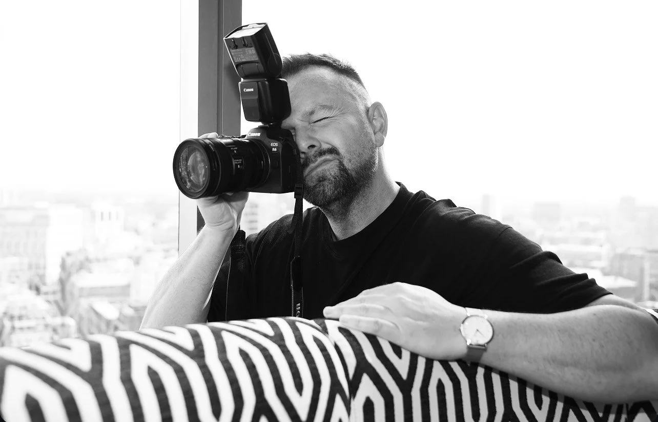 Man using a Canon EOS R6 camera with a flash, standing near a window, with a cityscape in the background and a patterned fabric in the foreground.