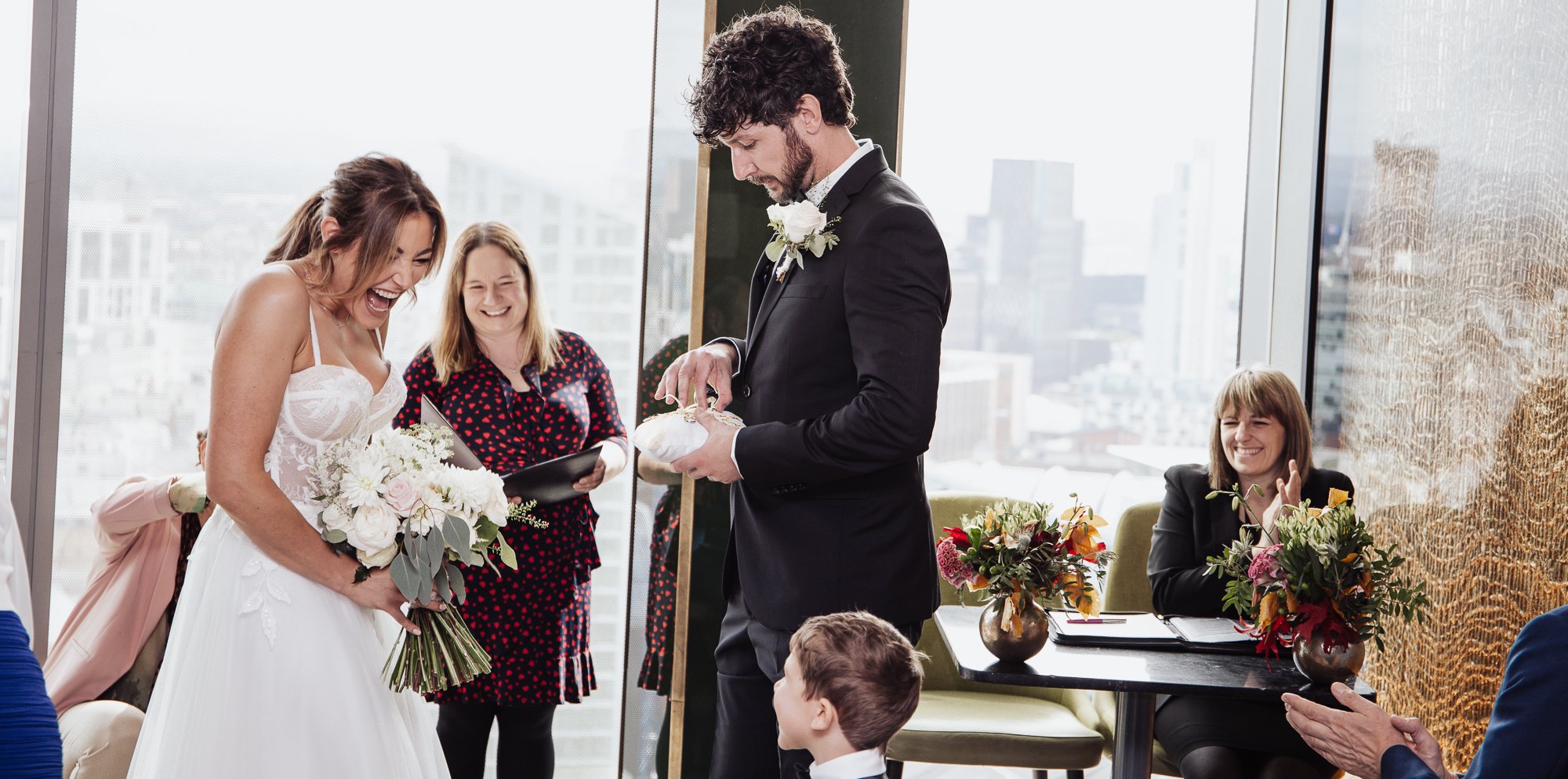 Bride in a white dress holding a bouquet laughing, groom in a suit looking at a ring pillow, indoors with guests seated and a city view.