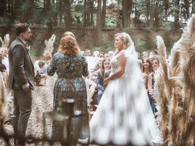 Outdoor wedding ceremony in a forest with a bride, groom, and officiant. Guests are seated on either side, surrounded by tall grass decorations.