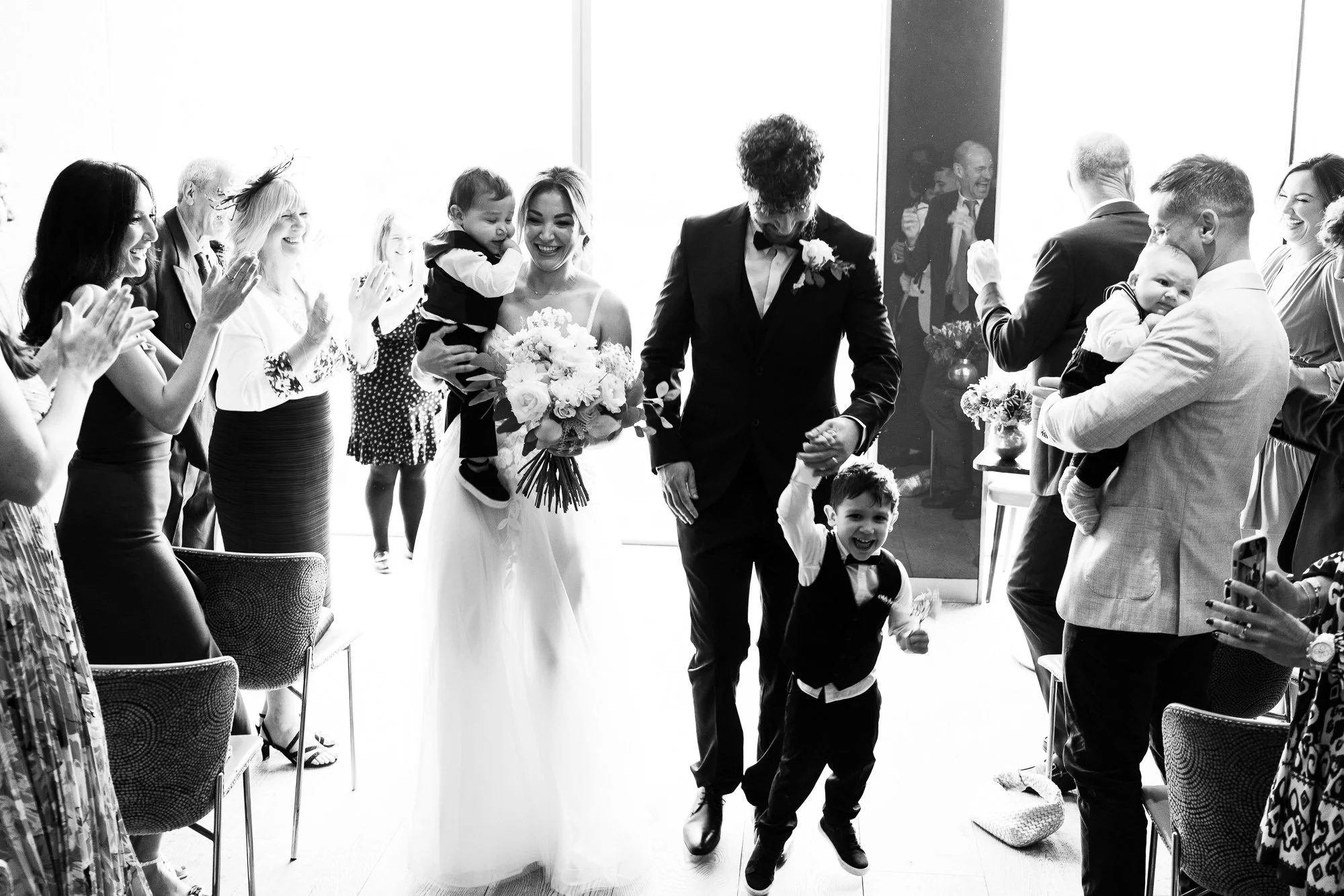 A bride and groom walking down the aisle with children, while guests clap and celebrate.