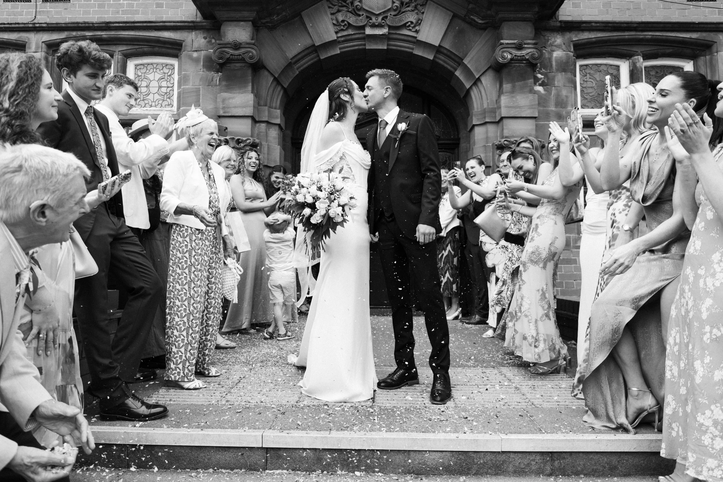 A black and white photo of a wedding ceremony outside a church. The bride and groom are kissing in the center, surrounded by friends and family who are clapping, smiling, and taking photos.
