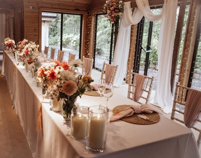 Elegant wedding reception table with floral centerpieces, candles, and white draped curtains in a rustic wooden venue.