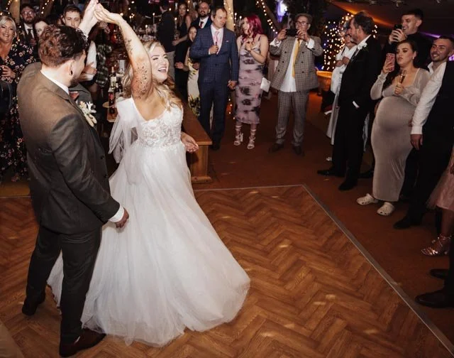 Bride and groom dancing in wedding attire surrounded by guests in a decorated indoor venue.