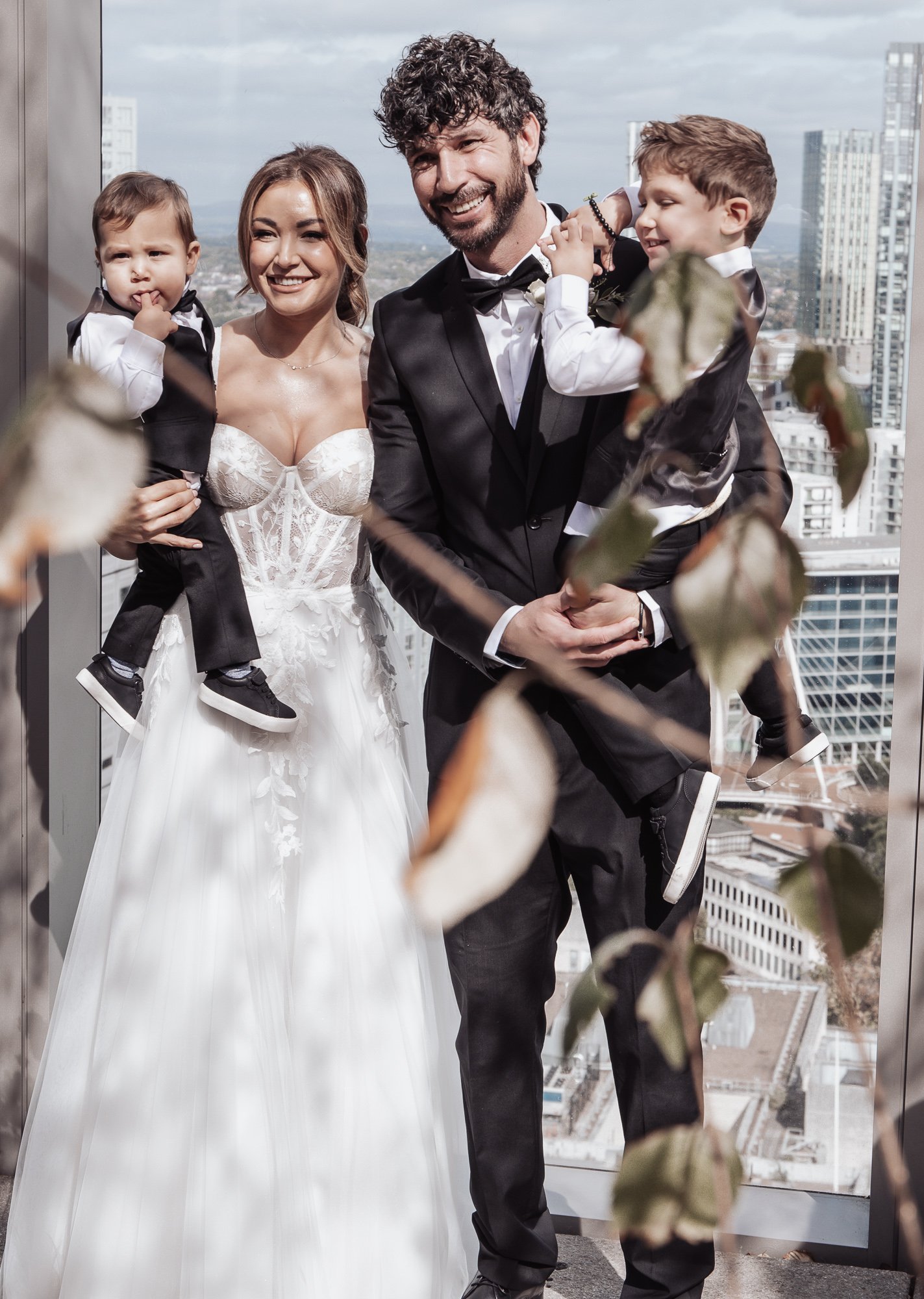A smiling couple dressed in wedding attire, standing with two young children in similar formal wear, with a cityscape in the background.