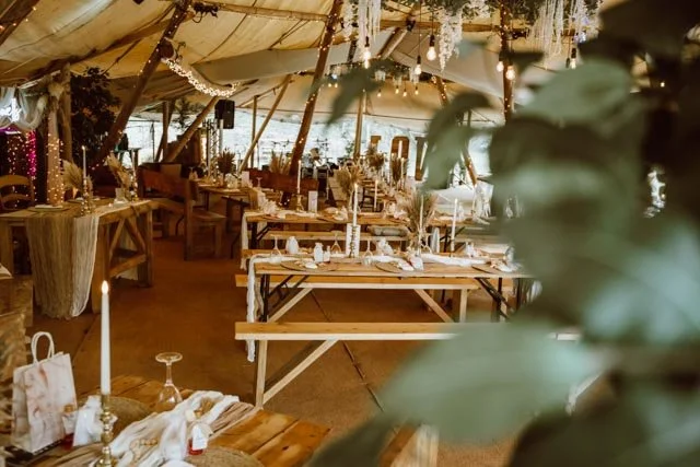 Rustic tent wedding reception with wooden tables, benches, hanging lights, and decorative foliage.