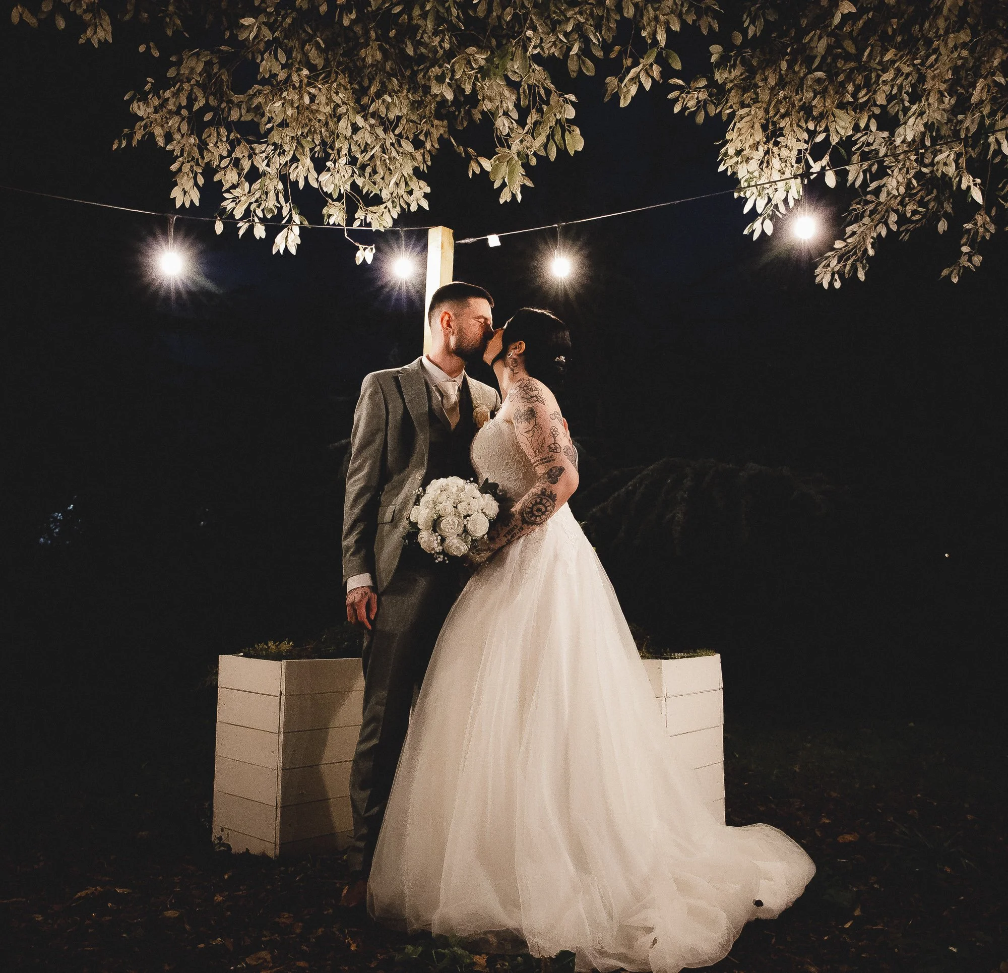 Bride and groom kissing under string lights at night, she is holding a bouquet, both standing by a tree.