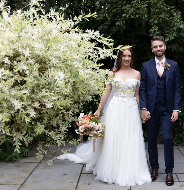 A bride and groom holding hands, standing outdoors beside green foliage. The bride is wearing a white wedding dress and holding a colorful bouquet. The groom is in a dark suit with a tie.