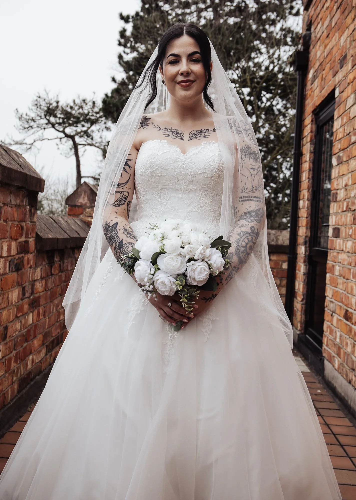 Bride in white lace gown holding bouquet of white flowers, with tattoos on arms, standing outdoors near brick walls.
