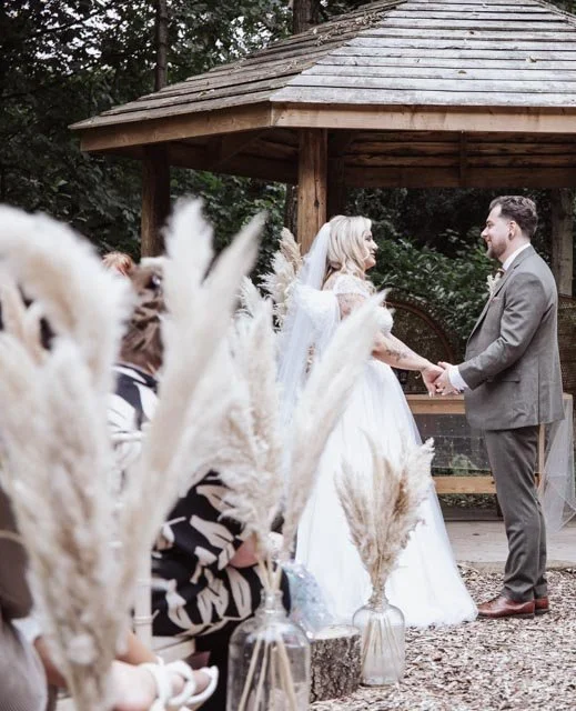 Bride and groom exchanging vows outdoors under wooden gazebo, decorated with pampas grass in glass vases, surrounded by guests.