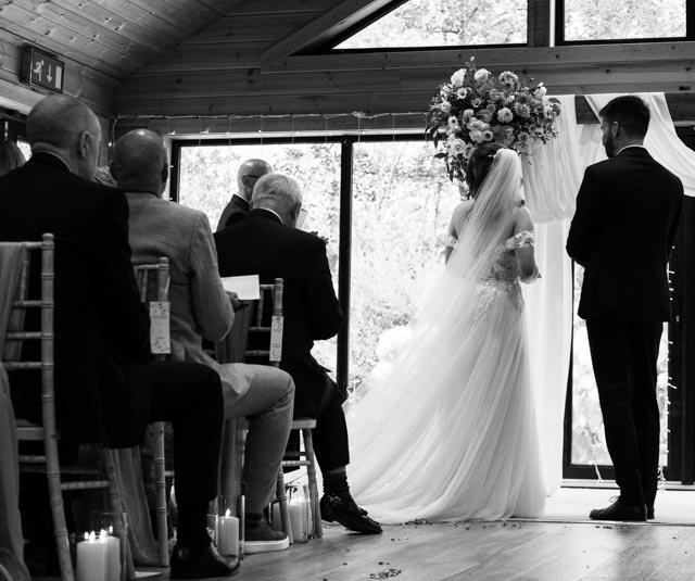 Black and white photo of a wedding ceremony with a bride in a gown and a groom standing, facing a window. Seated guests watch the ceremony in a wooden venue.