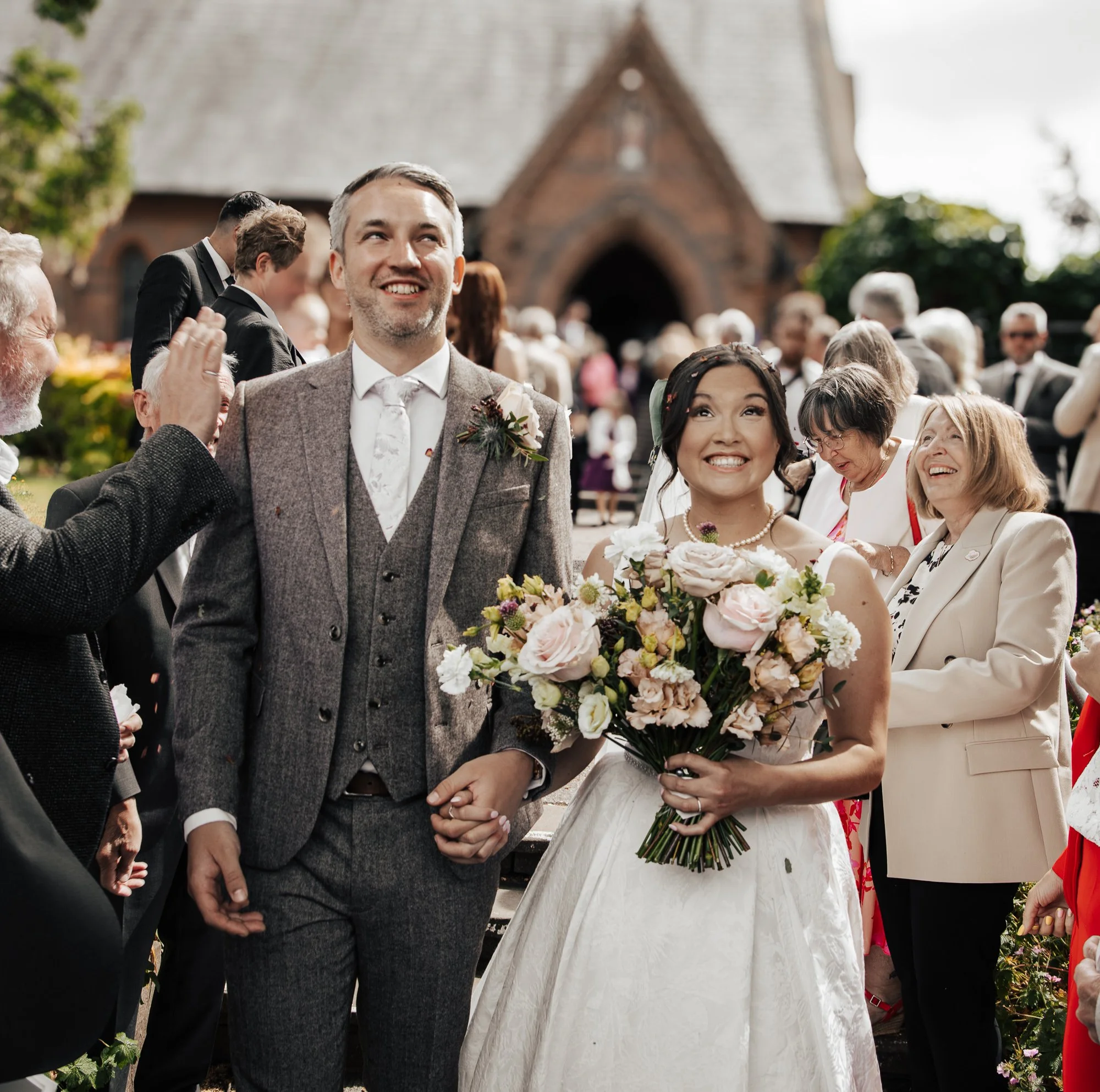 A bride and groom holding hands and smiling at their wedding, surrounded by guests outside a church, with the bride holding a large bouquet of flowers.