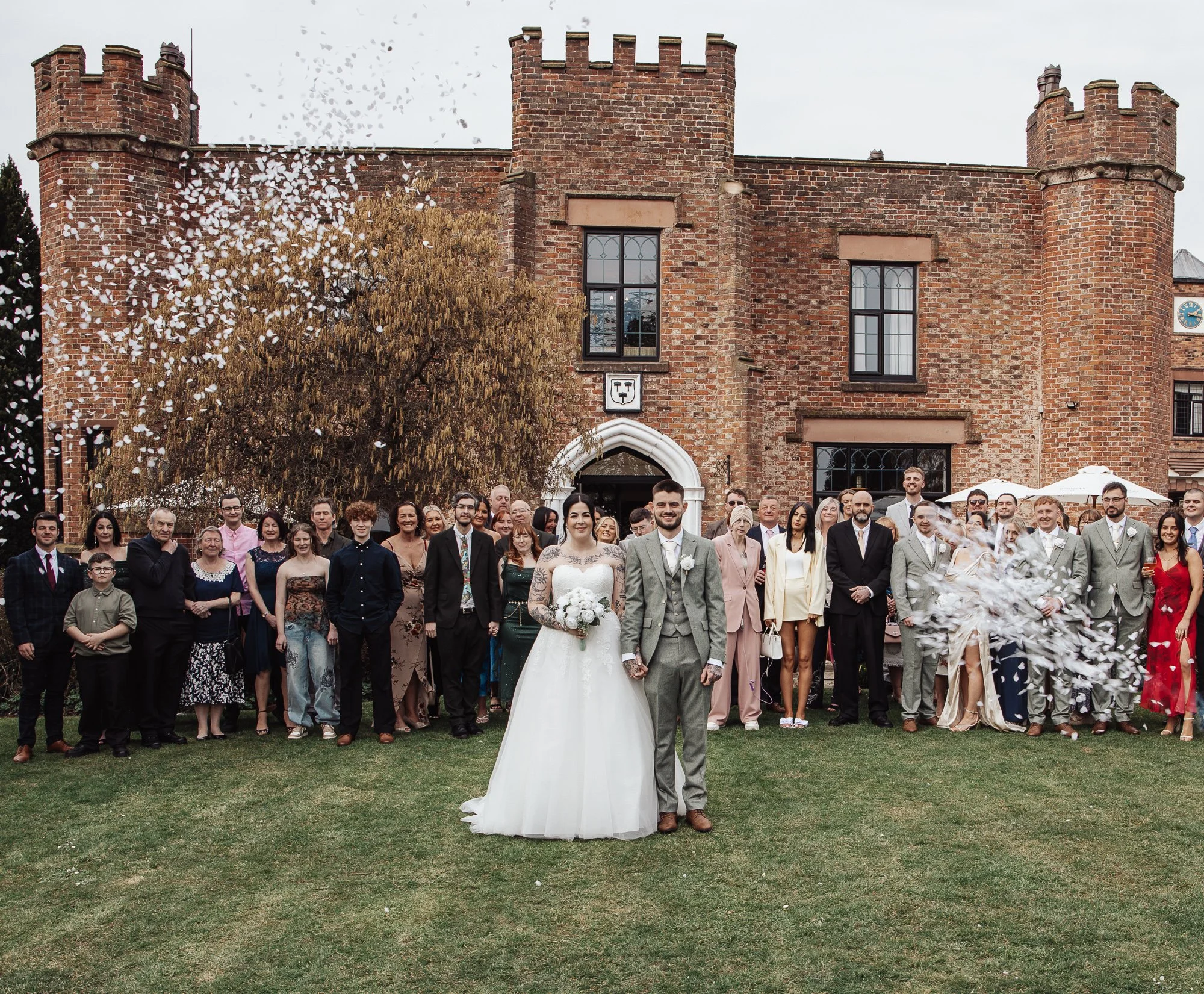 A wedding party stands in front of a brick building. The bride and groom are centered, surrounded by guests. The bride is in a white gown, and the groom wears a light gray suit. Flower petals are being thrown in the air. The building has a castle-like appearance with turrets.