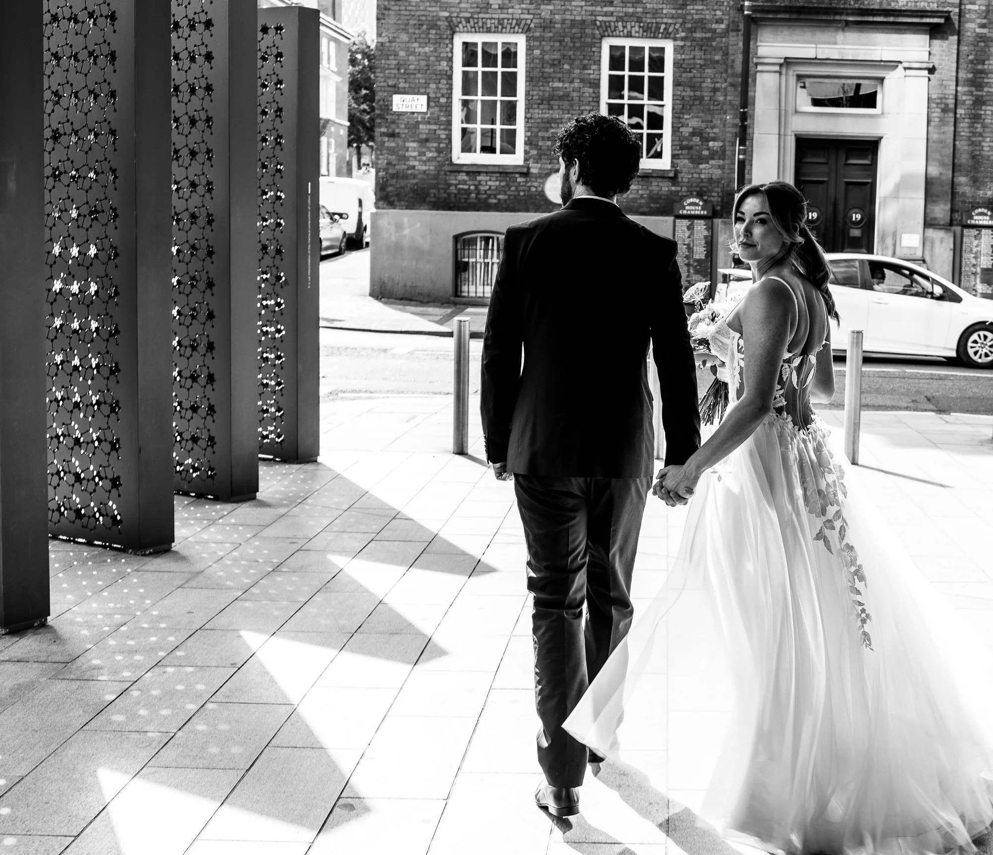 A black and white photo of a bride and groom walking hand in hand along a city street. The bride is wearing a wedding gown, and the groom is in a suit. They are outside near patterned columns, with a car and a building visible in the background.