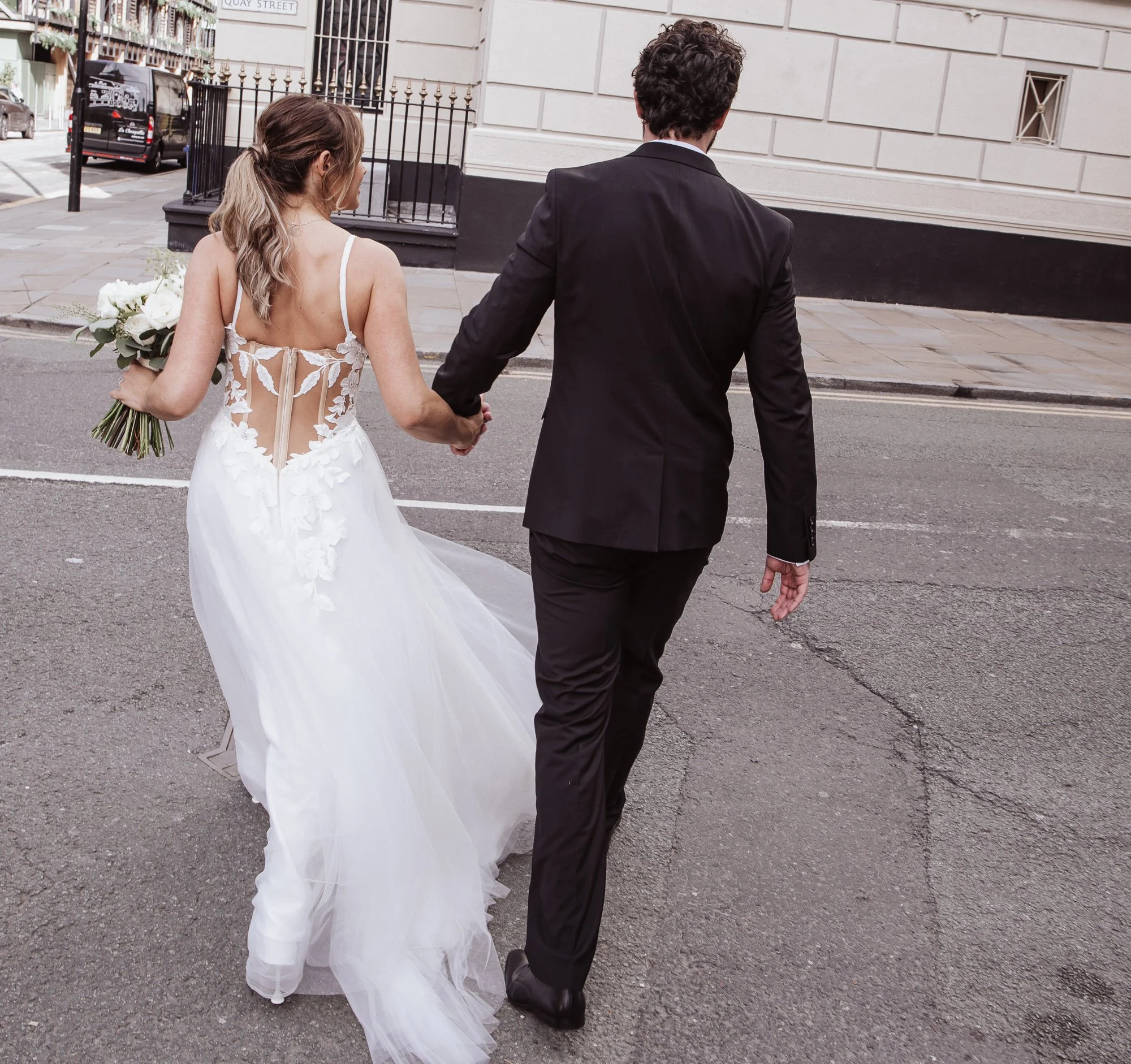 Bride in a white gown holding a bouquet, walking hand in hand with a groom in a black suit on a street.