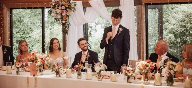 Wedding reception with decorated table, bridesmaid, bride, groom, best man giving a speech, and family seated, surrounded by floral arrangements.