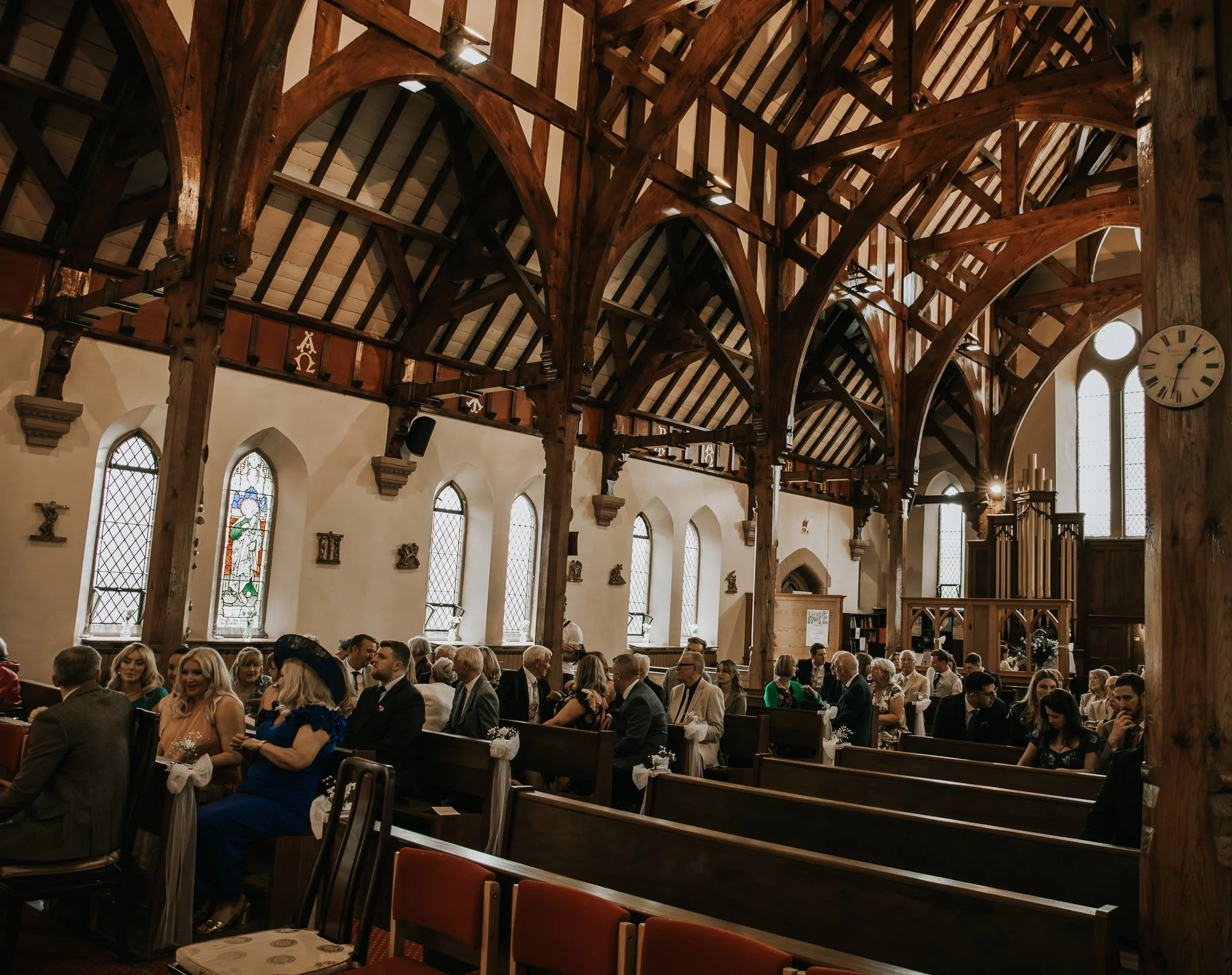 People seated in pews inside a church during a wedding or ceremony with stained glass windows and wooden beams.